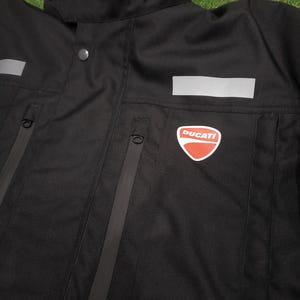 May include: Black motorcycle jacket with the word "DUCATI" in red on the collar. The jacket has a red and white Ducati logo on the chest, reflective panels, and zippered pockets. The jacket is made of a durable, weather-resistant material.