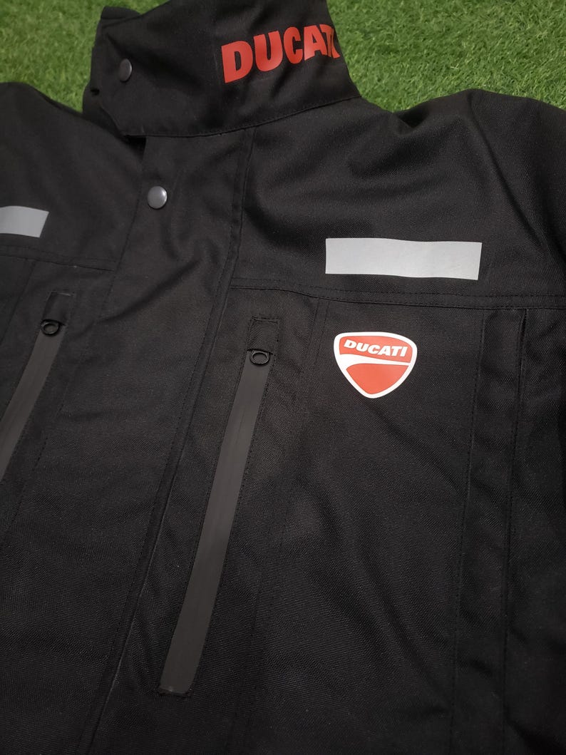 May include: Black motorcycle jacket with the word "DUCATI" in red on the collar. The jacket has a red and white Ducati logo on the chest, reflective panels, and zippered pockets. The jacket is made of a durable, weather-resistant material.