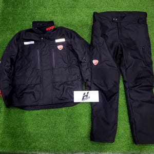 May include: Black motorcycle jacket and pants set. The jacket has the word "DUCATI" in red on the collar and sleeves, and a red logo on the chest. The pants also have a red logo. The set is displayed on a green surface.