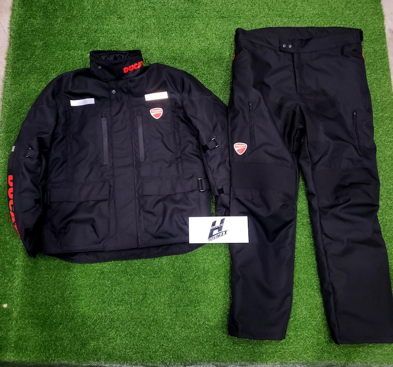 May include: Black motorcycle jacket and pants set. The jacket has the word "DUCATI" in red on the collar and sleeves, and a red logo on the chest. The pants also have a red logo. The set is displayed on a green surface.