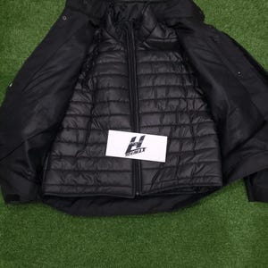 May include: Black jacket with a quilted inner layer, unzipped to reveal the interior. The outer shell has a collar and button closures. A white rectangular tag with the text "HOSIMAX" is visible.