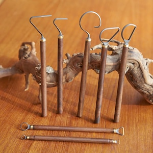 May include: A set of six metal and wood tools with different shaped ends. The tools have dark brown wooden handles with brass-coloured accents. The tools are displayed on a wooden surface, with a piece of driftwood in the background.