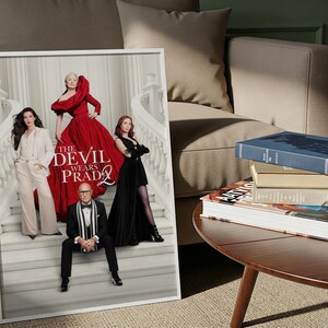 May include: A framed poster with the text "The Devil Wears Prada 2" features four people in stylish attire. A stack of books sits on a round wooden table next to a beige sofa. The scene is set in a well-lit room.