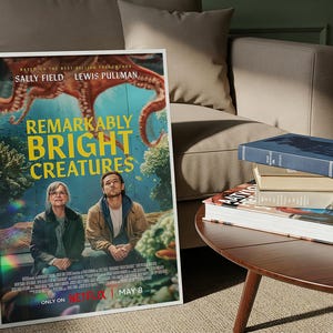 May include: A framed poster for "Remarkably Bright Creatures" with Sally Field and Lewis Pullman. The poster has a blue and green background with an octopus illustration. Several books are stacked on a wooden table next to a beige sofa.