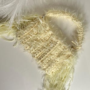 Puede incluir: Un bolso de mano color crema con un cuerpo tejido y un asa en bucle. El bolso está adornado con largas y esponjosas plumas que caen desde la parte inferior. La textura del bolso es una mezcla de elementos tejidos y plumosos.