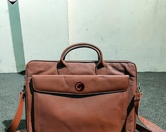 Puede incluir: Un maletín de cuero marrón con asa superior y bandolera desmontable. El maletín tiene un bolsillo frontal y un logotipo en la parte delantera. La bolsa es de cuero texturizado y tiene forma rectangular.