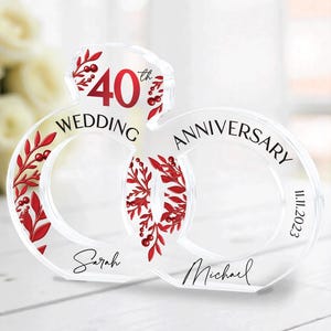 May include: Clear acrylic wedding anniversary ring decoration. Features the text "40th WEDDING ANNIVERSARY" with the names "Sarah" and "Michael" and the date "11.11.2023". Red floral accents adorn the rings.