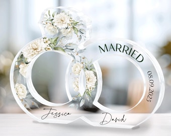 Peut inclure: Décoration de mariage en acrylique transparent en forme d'anneaux entrelacés, ornée de motifs floraux. Le mot « Married » et la date « 08.09.2023 » sont imprimés. Les noms « Jessica » et « David » sont imprimés à la base.