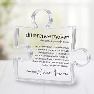 May include: A clear acrylic puzzle piece with the words "difference maker" printed in black. The definition of "difference maker" is included, along with the name "Emma Harris". The puzzle piece is on a white surface.