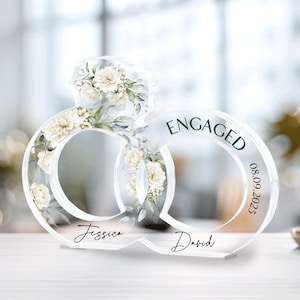 Könnte beinhalten: Transparente Acryl-Verlobungsring-Dekoration mit floralen Akzenten und dem Wort "ENGAGED". Das Datum "08.09.2025" ist auf dem Ring aufgedruckt, zusammen mit den Namen "Jessica" und "David".