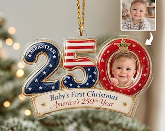 Peut inclure: Une décoration de Noël en acrylique transparent en forme de nombre 250, avec une section bleue pailletée à étoiles blanches, une section rayée rouge et blanche, et un cadre circulaire rouge contenant une photo de bébé et l'année 2026. Texte : Baby's First Christmas America's 250th Year.