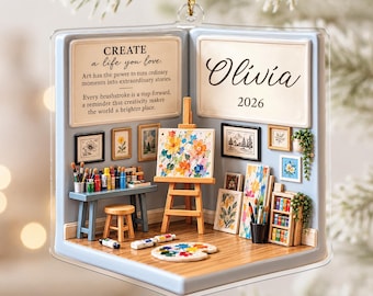 Peut inclure: Un ornement suspendu carré et transparent représentant un atelier d'artiste miniature. L'intérieur montre un bureau, un chevalet avec une peinture florale, un tabouret et des étagères. Texte : CREATE a life you love. Olivia 2026. Cordon doré.