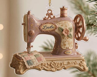 Peut inclure: Une décoration en forme de machine à coudre mauve et crème avec une bobine de fil dorée, une fleur en relief et un morceau de tissu matelassé. Le prénom Linda est inscrit en lettres cursives avec l'année 2026 près de la base.