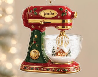 Peut inclure: Une décoration de Noël en forme de robot pâtissier rouge et vert avec des détails dorés, des feuilles de houx et le prénom Amelia inscrit sur le devant. Le bol mélangeur transparent contient une scène de maison en pain d'épices.