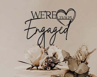 Peut inclure: Un gâteau de mariage blanc à trois étages décoré de compositions florales et d'un cake topper noir avec l'inscription "We're Engaged" avec un cœur et une date.