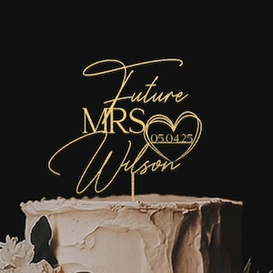 Può includere: Una torta nuziale a due piani con glassa bianca e decorazioni floreali. Un cake topper dorato recita "Future MRS Wilson 05.04.25" in elegante scrittura. La torta è adornata con fiori e verde pesca e bianchi, su sfondo scuro.