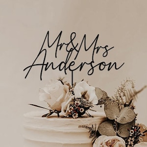 Peut inclure: Un gâteau de mariage blanc à trois niveaux, décoré de roses crème et de verdure. Le gâteau est surmonté d'un cake topper noir en écriture cursive qui dit "Mr & Mrs Anderson". Le gâteau a un glaçage au beurre texturé.