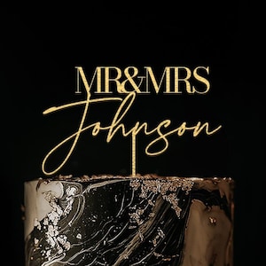 May include: A two-tiered wedding cake with a black and white marble design, adorned with gold leaf accents. A gold cake topper reads "MR & MRS Johnson" in elegant script, set against a dark background.