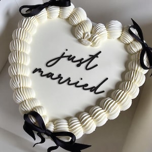 Puede incluir: Un pastel en forma de corazón con glaseado blanco y borde de crema de mantequilla. El pastel está decorado con lazos de cinta negra y las palabras "just married" en escritura negra. El pastel está en una caja blanca.