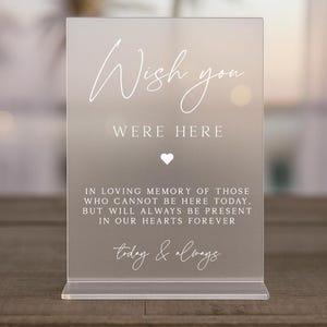 May include: A clear acrylic sign with the words "Wish you were here" in elegant script. Below, the text reads "In loving memory..." with a heart symbol. The sign is designed to stand on a table.
