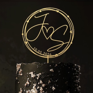 Może przedstawiać: Dwupoziomowe czarne ciasto ze złotymi akcentami z liści, zwieńczone złotym topperem. Topper przedstawia splecione inicjały "J" i "S" z sercem, otoczone ozdobną ramką oraz datą "10.05.2026".