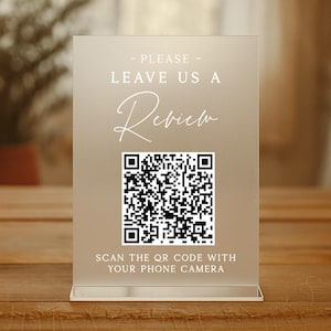 Könnte beinhalten: Ein klarer Acrylschild mit dem Text "- PLEASE - LEAVE US A Review" in weißer Schrift. Unter dem Text befindet sich ein schwarz-weißer QR-Code mit den Worten "SCAN THE QR CODE WITH YOUR PHONE CAMERA". Das Schild steht auf einer Holzoberfläche.
