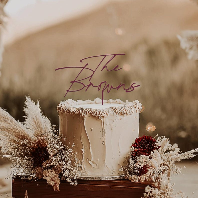 May include: A white frosted cake with a decorative edge, topped with a purple cake topper that reads "The Browns." The cake is decorated with dried flowers and greenery, including pampas grass and burgundy flowers. The cake sits on a wooden surface.