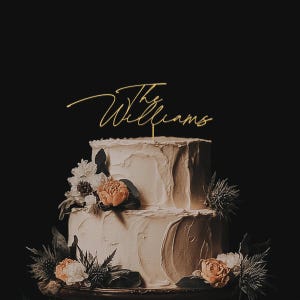 May include: A two-tiered wedding cake with white frosting and floral decorations. A gold-colored cake topper reads "The Williams" in cursive script. The cake is decorated with peach and white flowers and greenery.