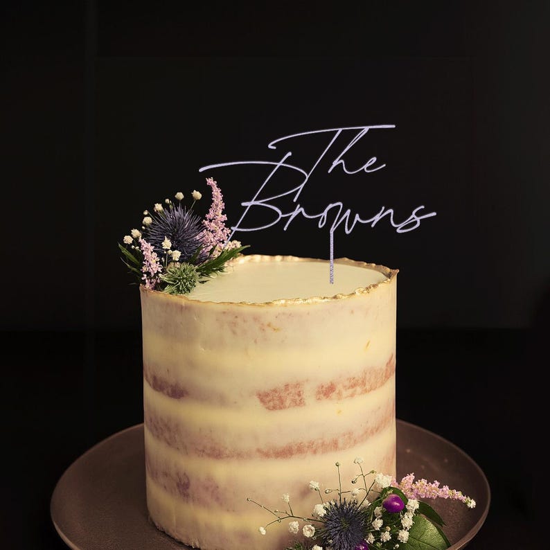 May include: A round, frosted cake with a gold-trimmed top, decorated with fresh flowers and a cake topper that reads "The Browns". The cake sits on a brown plate against a dark background. The cake is approximately 6 inches in diameter.