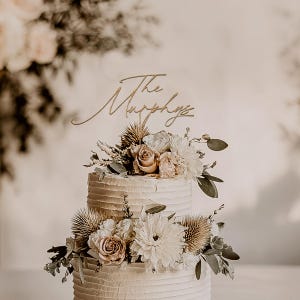May include: A two-tiered wedding cake with textured buttercream frosting, adorned with fresh flowers in shades of white, cream, and blush. A gold-colored cake topper reads "The Murphys". The cake is decorated with thistles and greenery.