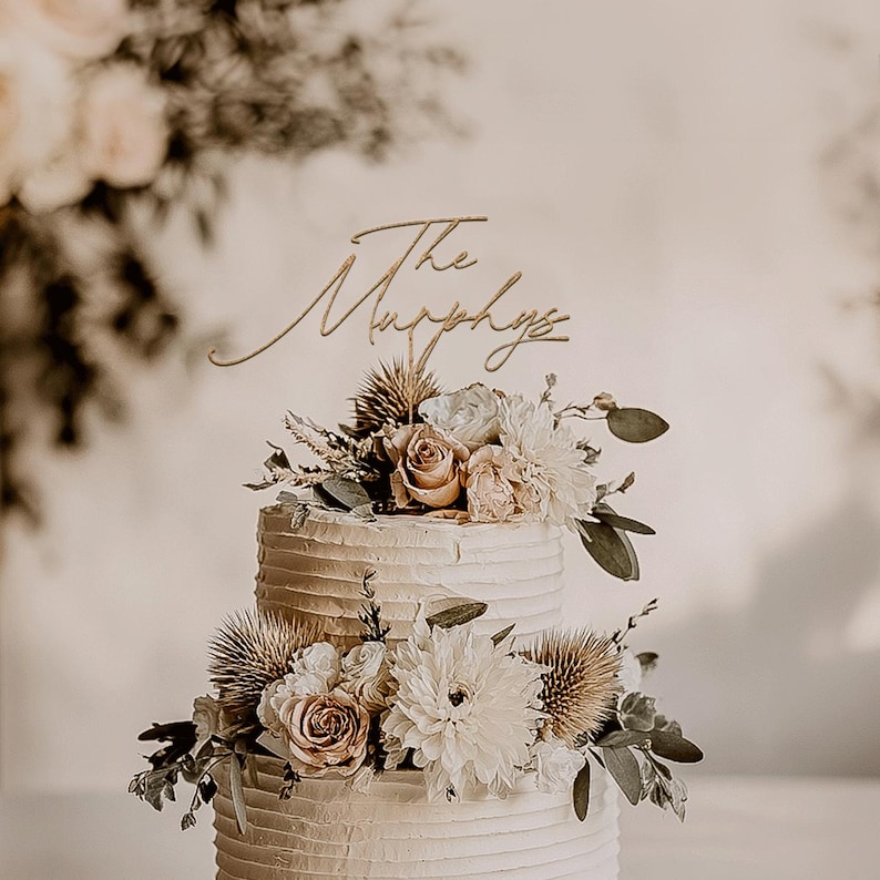 May include: A two-tiered wedding cake with textured buttercream frosting, adorned with fresh flowers in shades of white, cream, and blush. A gold-colored cake topper reads "The Murphys". The cake is decorated with thistles and greenery.