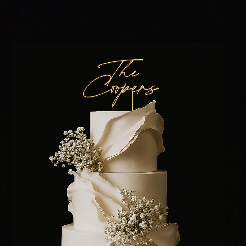 May include: A tiered white wedding cake with floral accents and a gold cake topper that reads "The Coopers" in elegant script. The cake has a smooth finish and is decorated with delicate white flowers.
