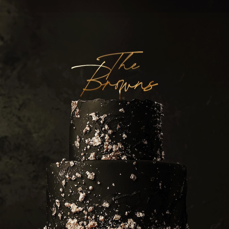 May include: A two-tiered black cake with gold leaf accents. A gold-colored cake topper reads "The Browns" in elegant script. The cake has a textured surface and is set against a dark background.