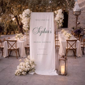 May include: A white fabric banner with the text "Welcome to Sophia's Bridal Shower" in a decorative font. The scene is set for a wedding reception, with tables, chairs, and floral decorations. A lantern with a candle is visible.