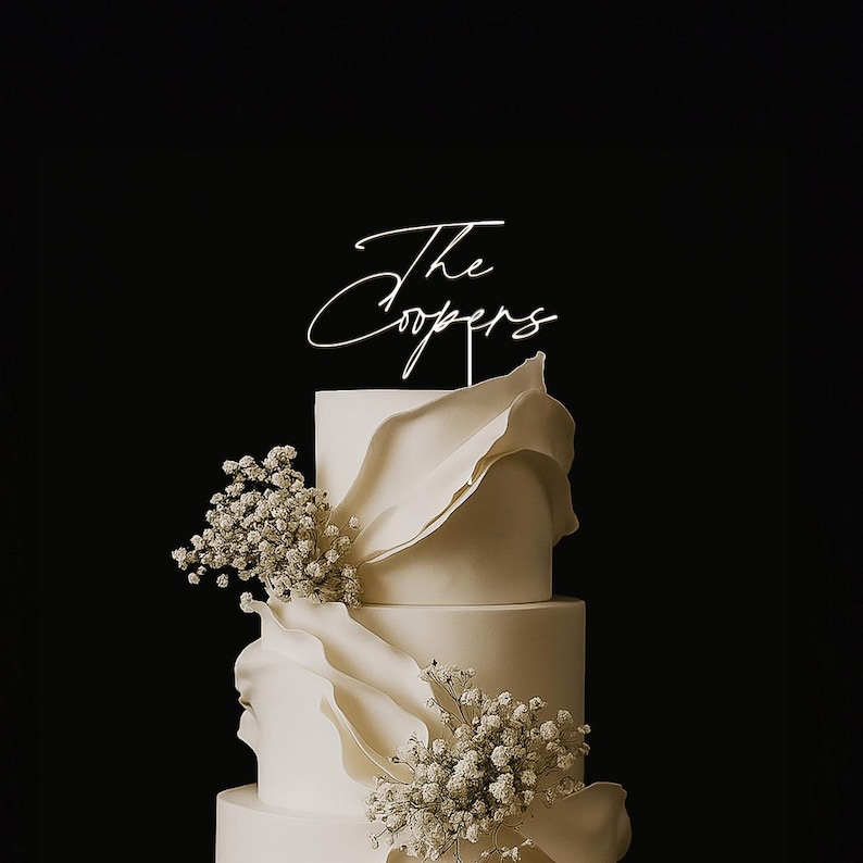 May include: A tiered white wedding cake with delicate floral accents and a topper that reads "The Coopers" in elegant script. The cake features soft, flowing decorations and is set against a dark background.