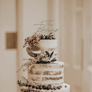 May include: A three-tiered wedding cake with a semi-naked design, decorated with fresh berries and greenery. A cake topper reads "The Browns" in elegant script. The cake is a centerpiece, perfect for a wedding or special event.