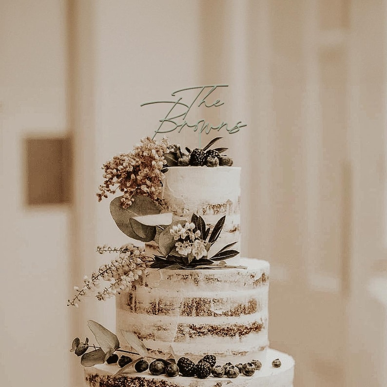 May include: A three-tiered wedding cake with a semi-naked design, decorated with fresh berries and greenery. A cake topper reads "The Browns" in elegant script. The cake is a centerpiece, perfect for a wedding or special event.