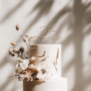 May include: A two-tiered white wedding cake with gold accents and floral decorations. The cake is topped with a gold-colored script that reads "The Adams". The cake is decorated with white flowers and dried foliage.