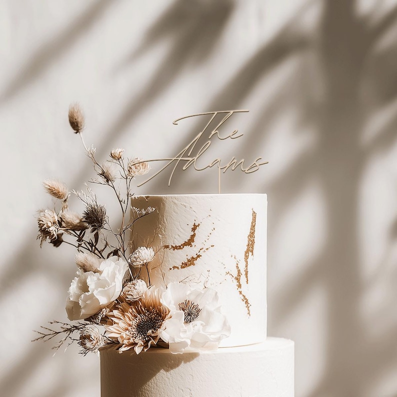 May include: A two-tiered white wedding cake with gold accents and floral decorations. The cake is topped with a gold-colored script that reads "The Adams". The cake is decorated with white flowers and dried foliage.