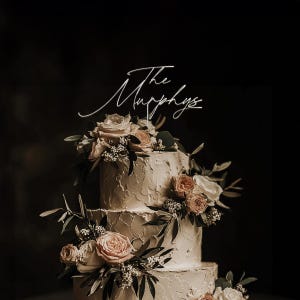 May include: A three-tiered wedding cake with textured buttercream frosting. The cake is decorated with fresh flowers in shades of pink and cream, and greenery. A white cake topper reads "The Murphys" in elegant script.