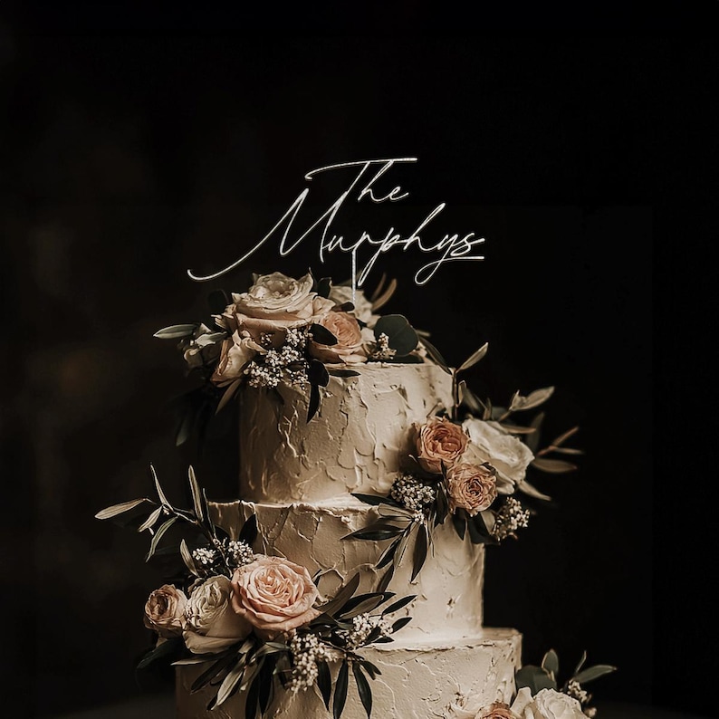 May include: A three-tiered wedding cake with textured buttercream frosting. The cake is decorated with fresh flowers in shades of pink and cream, and greenery. A white cake topper reads "The Murphys" in elegant script.