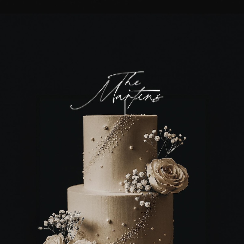 May include: A two-tiered wedding cake with a silver cake topper that reads "The Martins". The cake is decorated with white pearls, white flowers, and a large rose. The background is black.