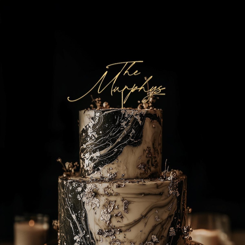 May include: A two-tiered wedding cake with a gold cake topper that reads "The Murphys." The cake has a marble design with black, white, and gold accents. Gold leaf and small floral decorations adorn the cake.