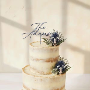 May include: A two-tiered wedding cake with a rustic design. The cake is decorated with fresh flowers and a navy blue cake topper that reads "The Adams". The cake has a cream-colored frosting and a golden edge.