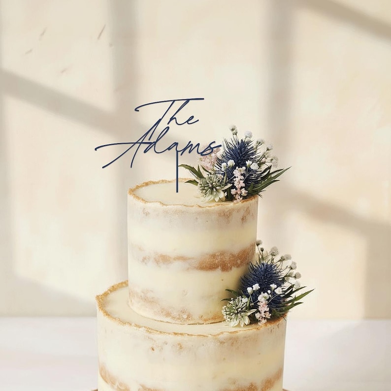 May include: A two-tiered wedding cake with a rustic design. The cake is decorated with fresh flowers and a navy blue cake topper that reads "The Adams". The cake has a cream-colored frosting and a golden edge.