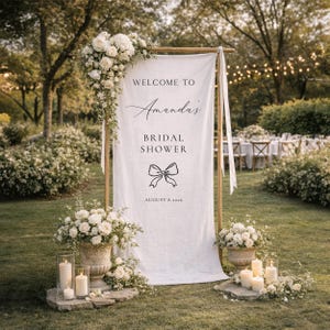 May include: A white fabric sign with the text "WELCOME TO Amanda's BRIDAL SHOWER" and a bow graphic, framed by a wooden structure. The sign is decorated with white flowers and candles, set in a garden setting.