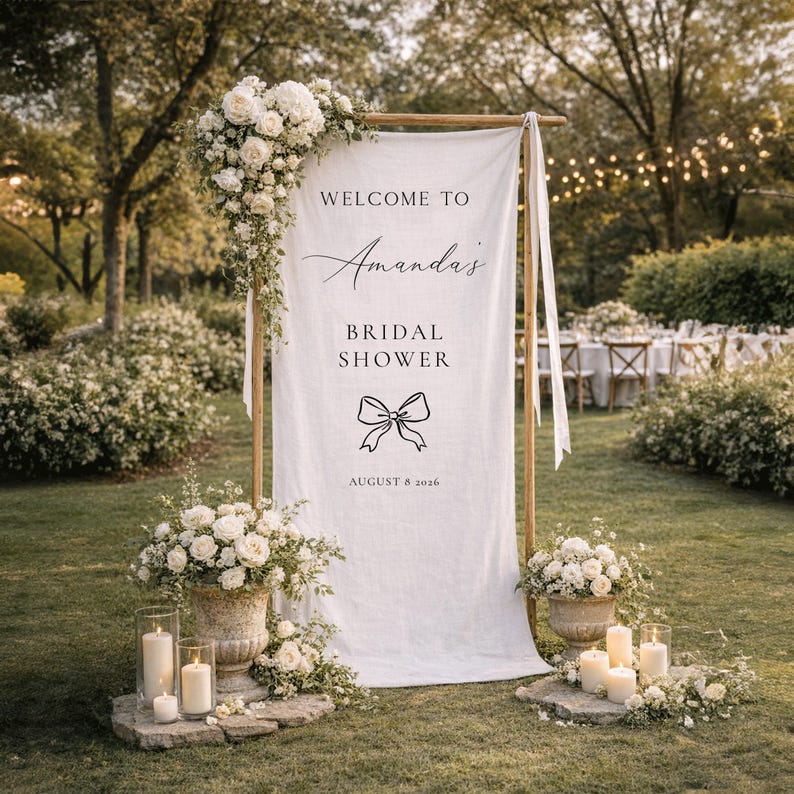May include: A white fabric sign with the text "WELCOME TO Amanda's BRIDAL SHOWER" and a bow graphic, framed by a wooden structure. The sign is decorated with white flowers and candles, set in a garden setting.