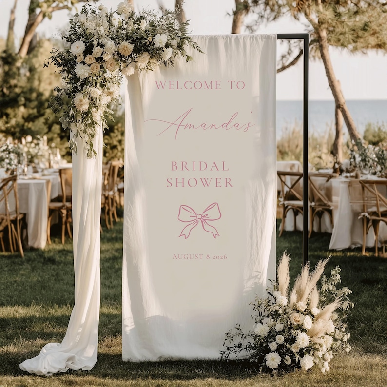 May include: A cream fabric sign with pink text reading "WELCOME TO Amanda's BRIDAL SHOWER" and a bow graphic. The date "AUGUST 8 2026" is also displayed. The sign is part of a wedding reception, decorated with floral arrangements.