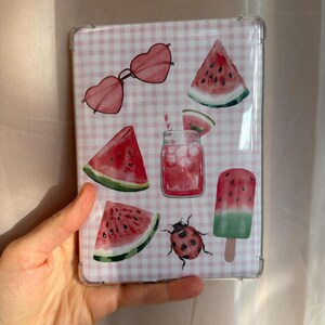 Pode incluir: Uma capa transparente para tablet com um padrão de fundo xadrez vichy em rosa claro e branco, apresentando várias ilustrações, incluindo óculos de sol em forma de coração, fatias de melancia, um frasco de vidro com bebida rosa e palhinha, uma joaninha e um gelado de melancia.