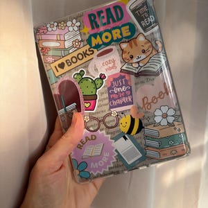 Peut inclure: Une coque de protection transparente pour tablette ou liseuse ornée de divers autocollants représentant des piles de livres, un chat qui lit, un cactus, une abeille et les textes READ MORE, I LOVE BOOKS, TIME TO READ, COZY VIBES et JUST ONE MORE CHAPTER.